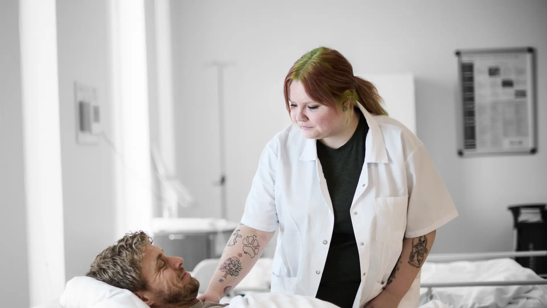 Social- og sundhedsassistent står ved hospitalsseng, hvor der ligger en anden elev i