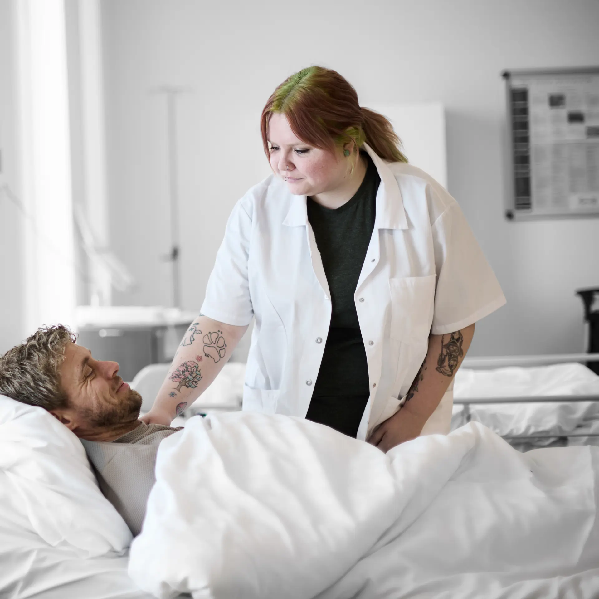 Social- og sundhedsassistent står ved hospitalsseng, hvor der ligger en anden elev i