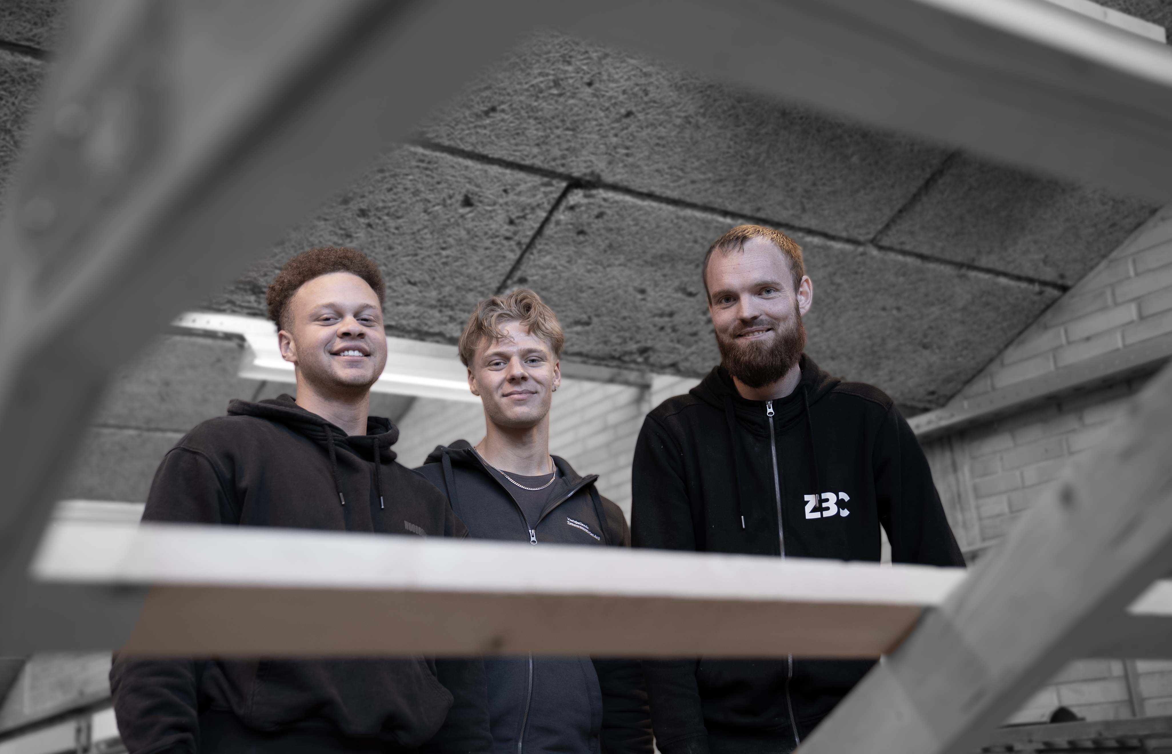 Tømrerelever på værksted bag træ smiler til kamera