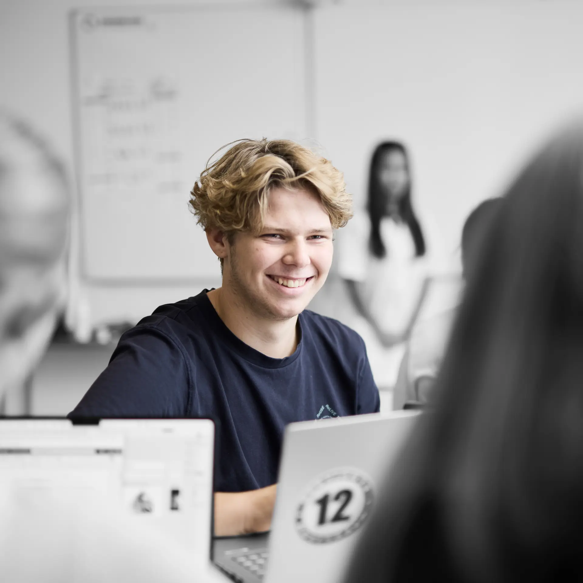 Pædagogisk assistent sidder ved et bord og smiler, mens han skal løse en opgave på computeren