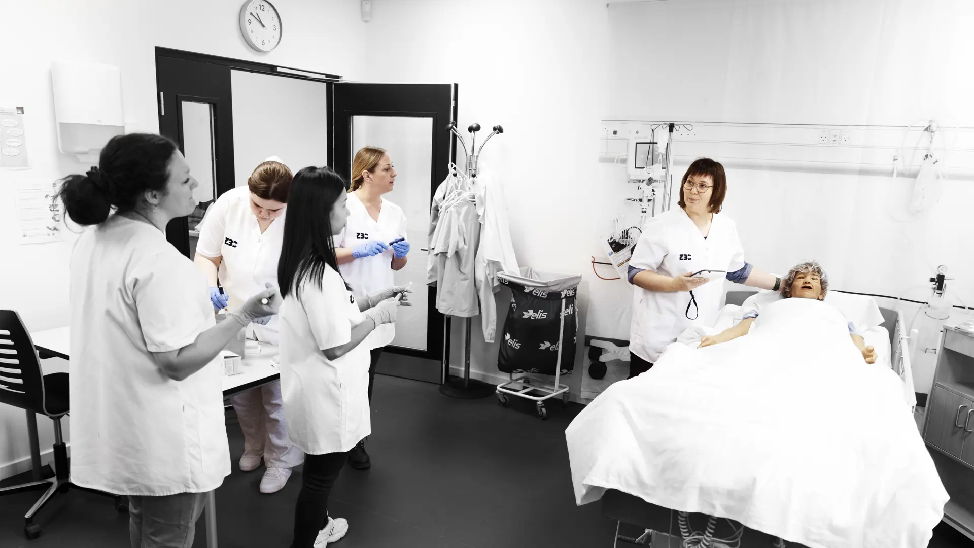 Social og sundhedsassistenter på demostue, hvor faglærere underviser ved dukke, som ligger i en hospitalseng