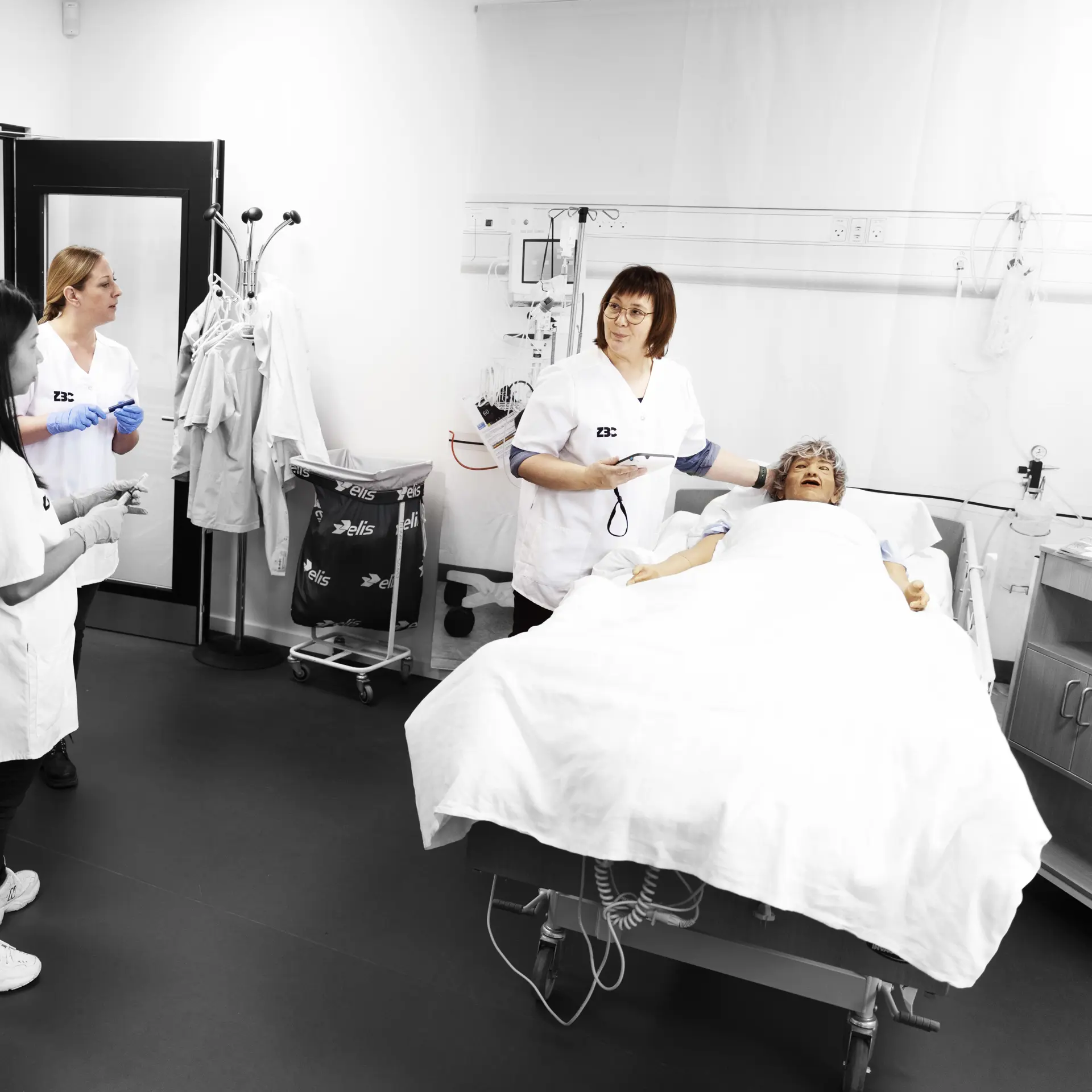 Social og sundhedsassistenter på demostue, hvor faglærere underviser ved dukke, som ligger i en hospitalseng