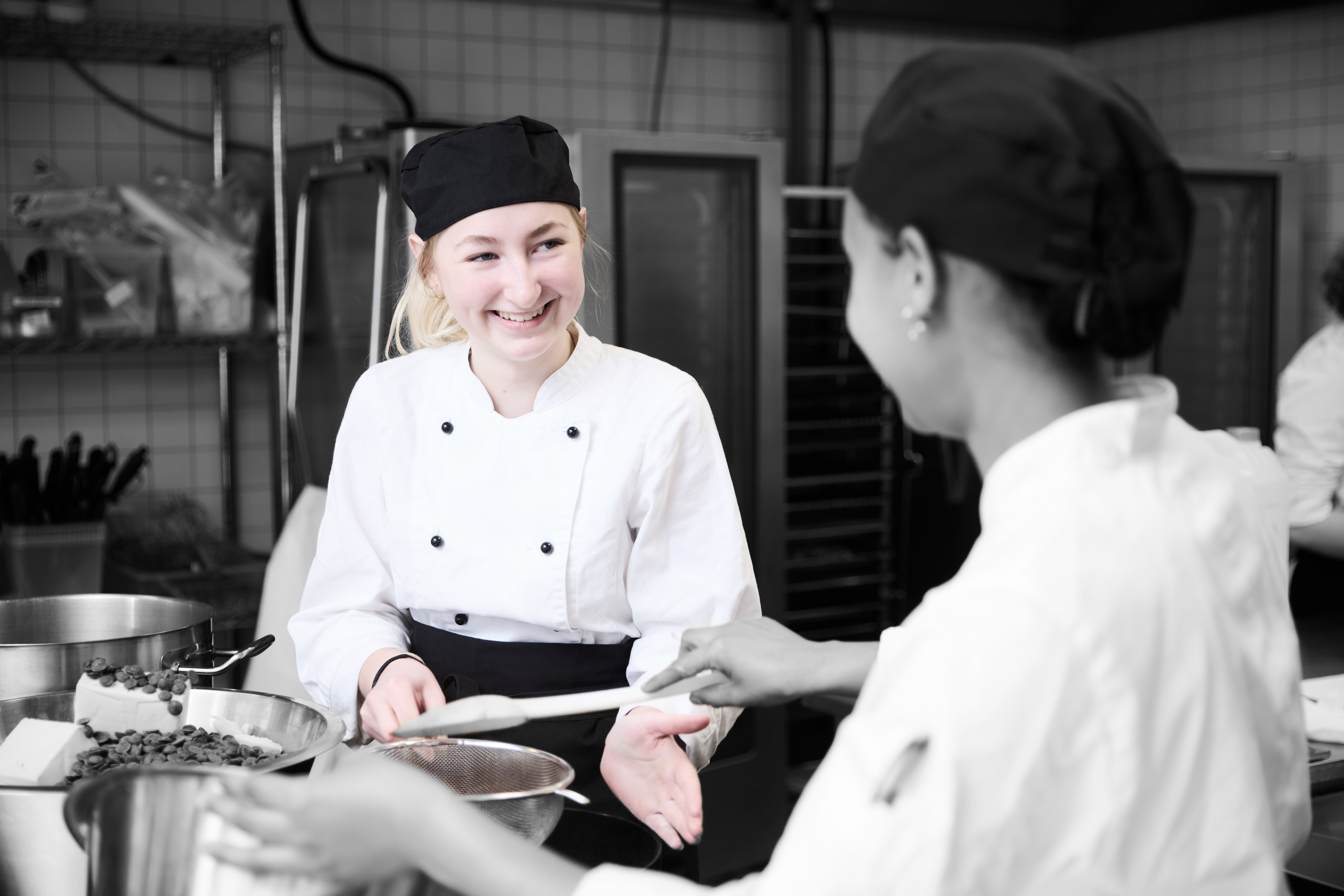 Ernæringsassistent i køkken smiler til elev, mens hun temperer chokolade